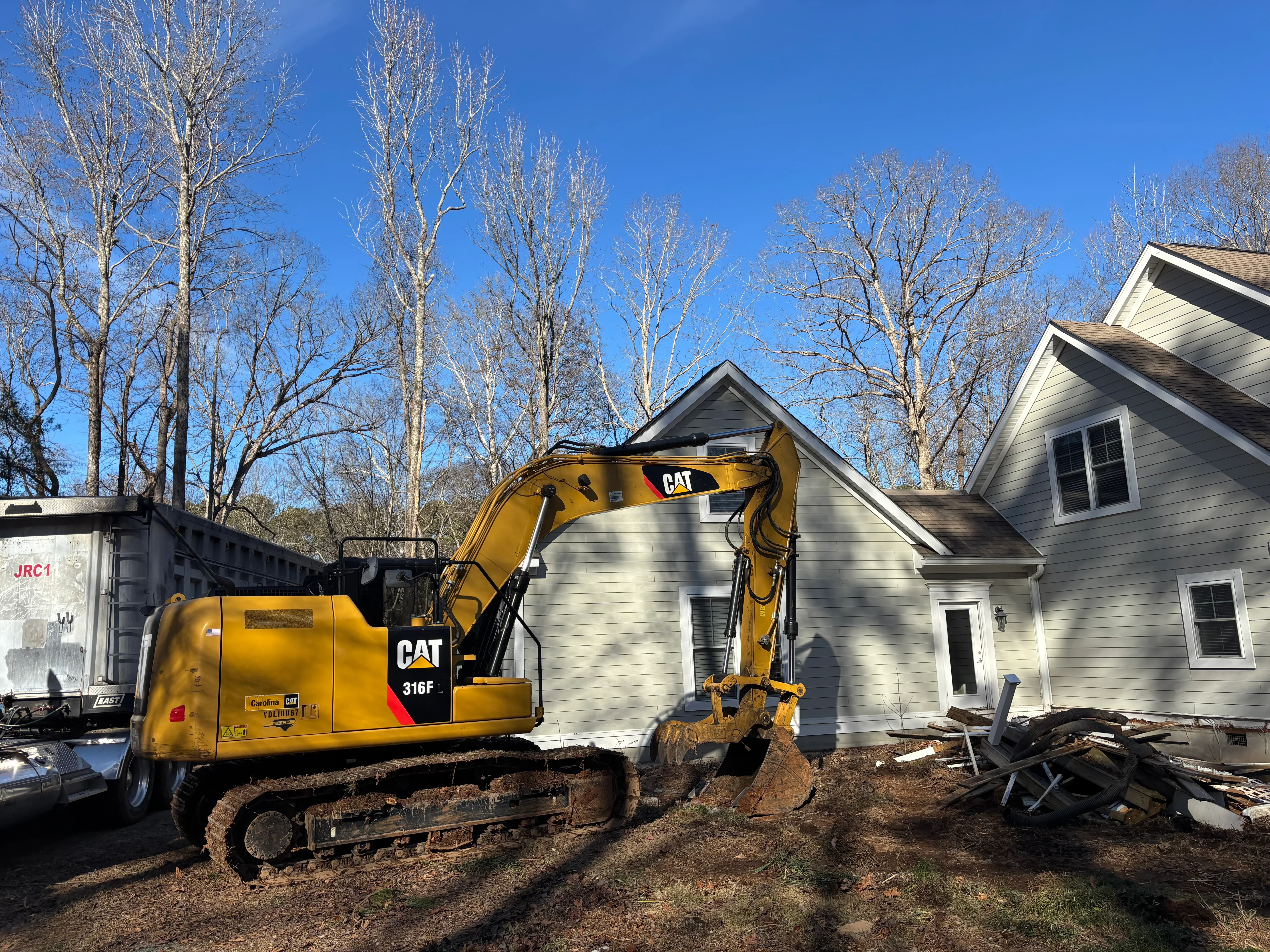 CAT 316F excavator digging foundation area next to white residential house with bare trees