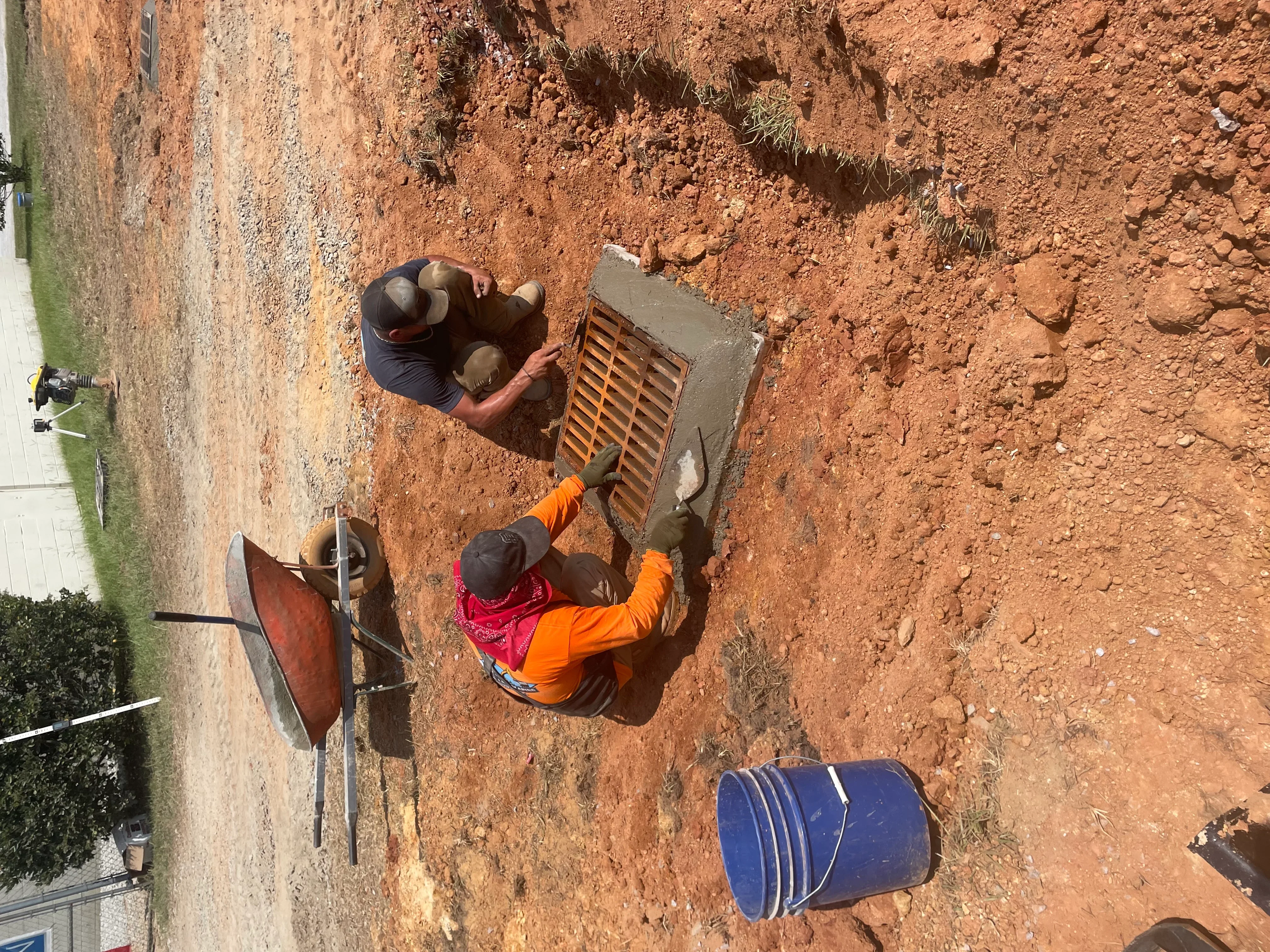 Workers installing metal drainage grate inlet box in excavated clay soil with grading work