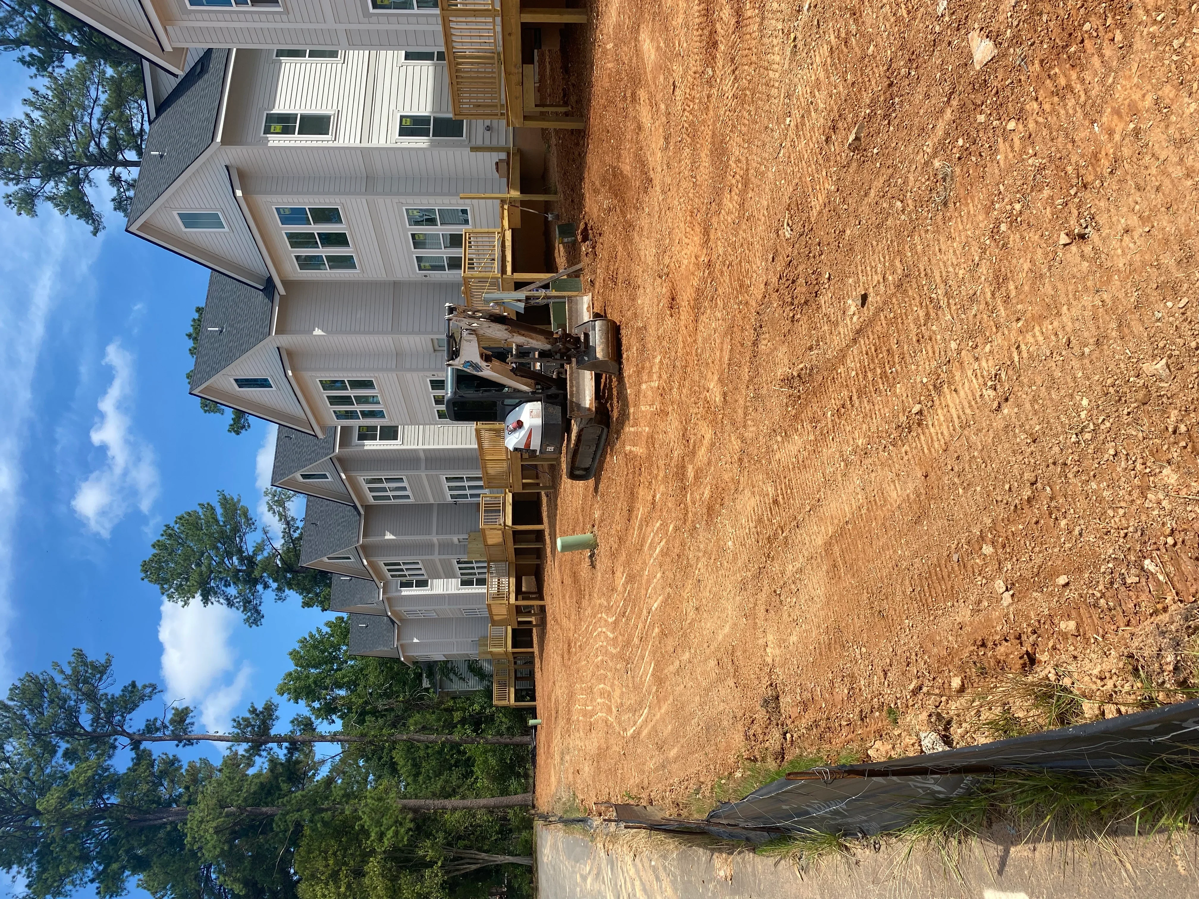 Excavated residential lot with graded soil, metal-sided houses under construction, and wooden deck framing