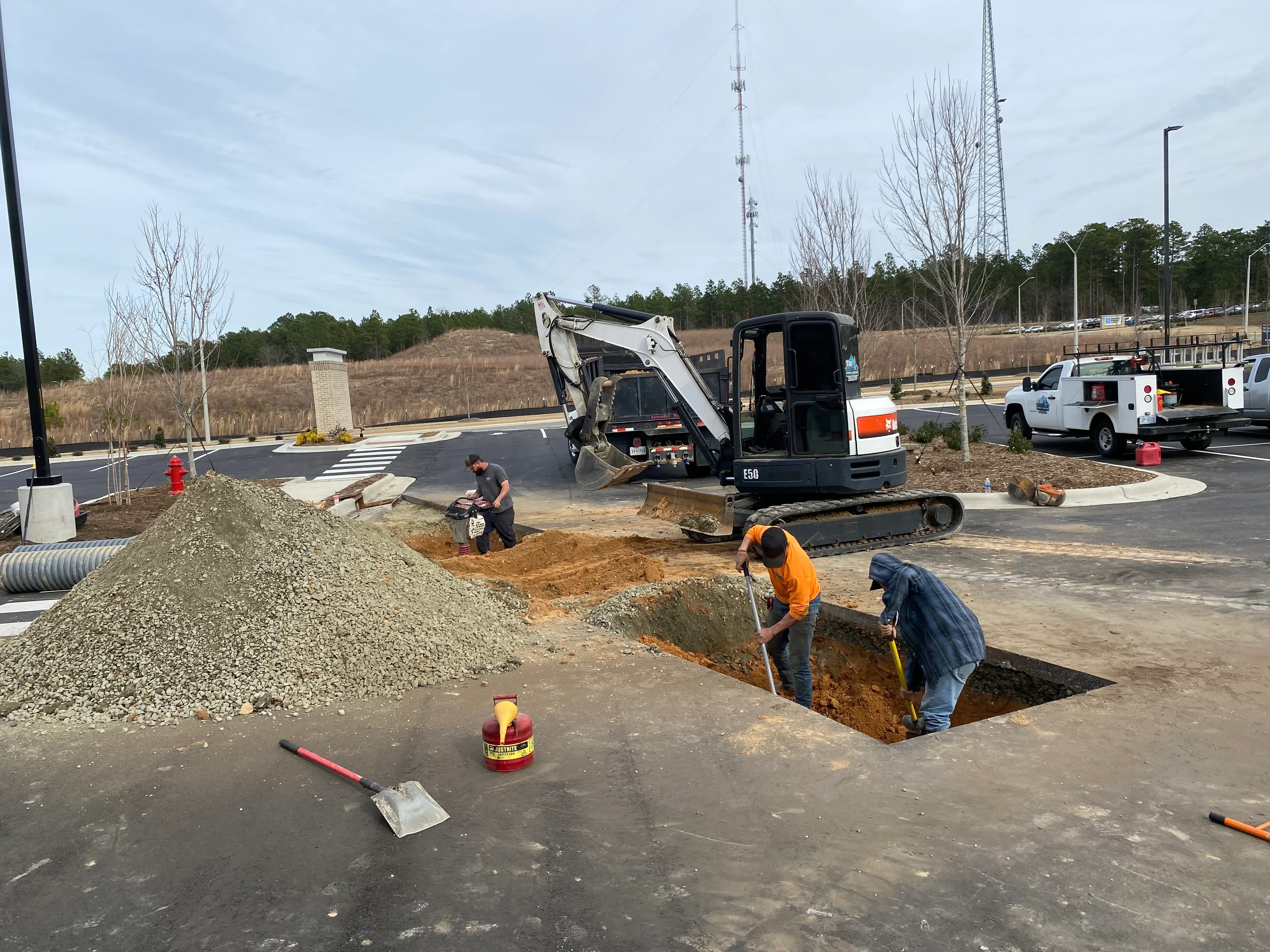 Excavator digging utility trench at commercial site with workers and gravel stockpile