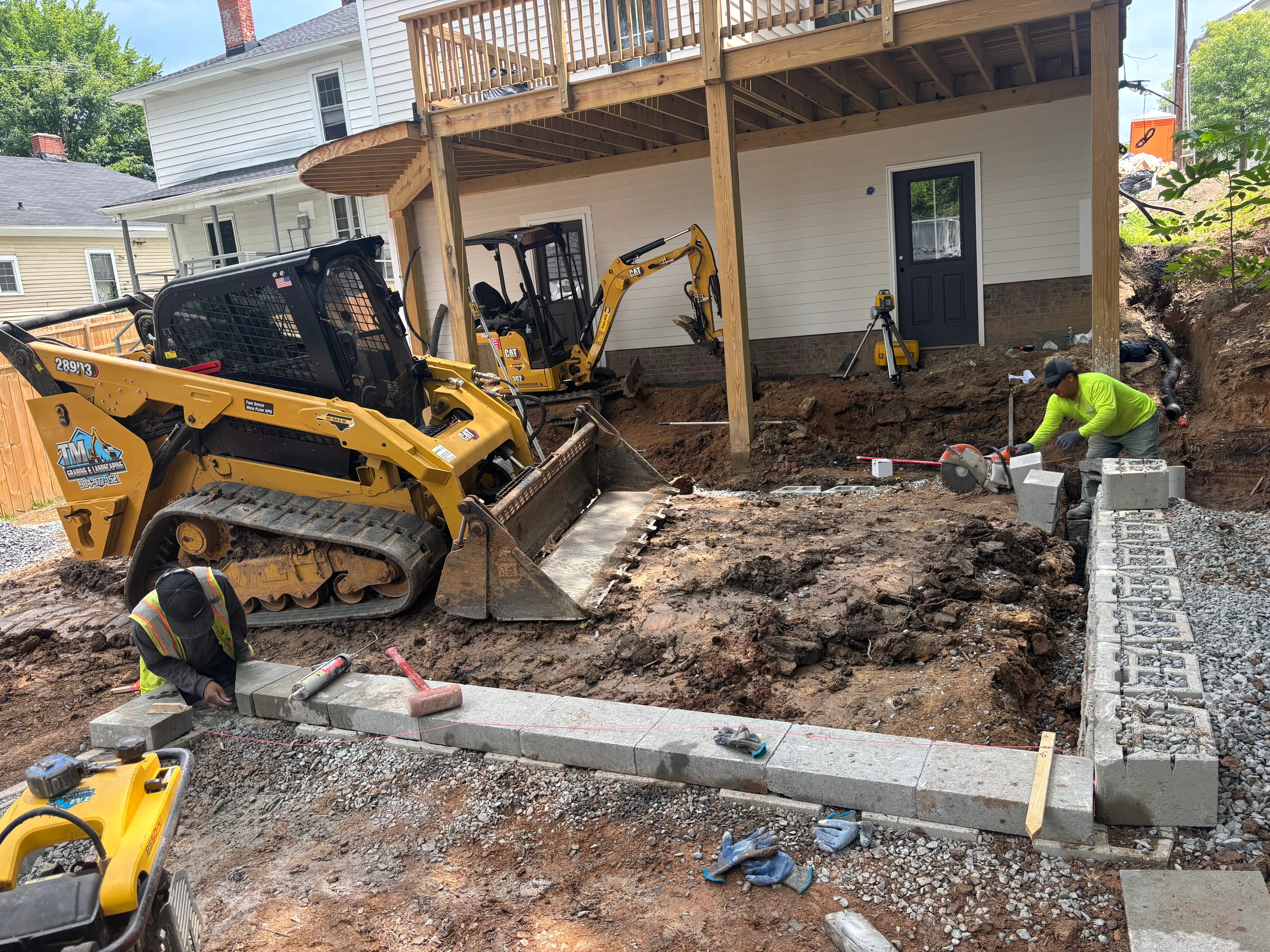 Excavation site with skid steer loader, concrete retaining wall blocks, and graded soil foundation