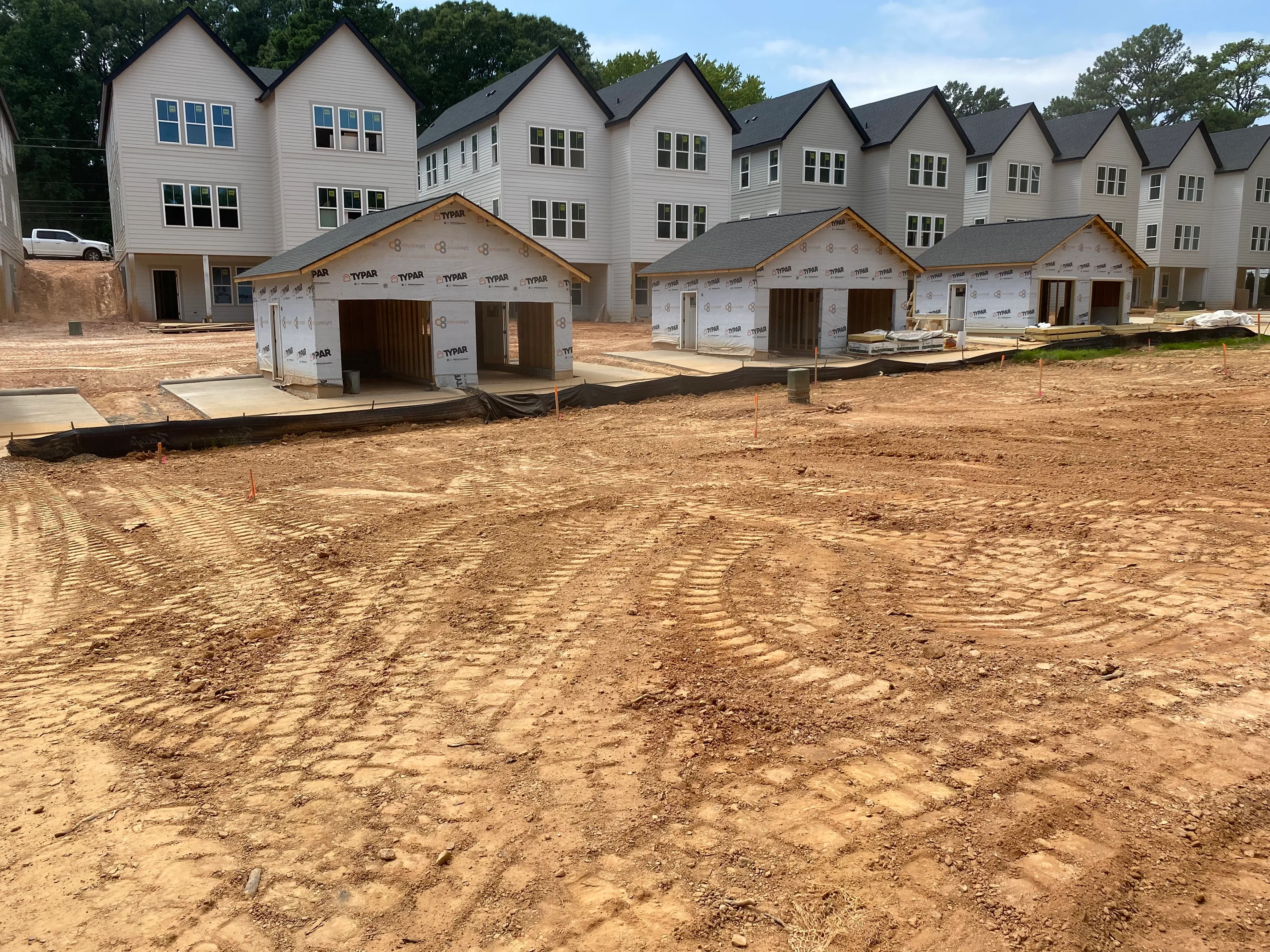Graded dirt site with concrete pads and framed townhomes under construction in background