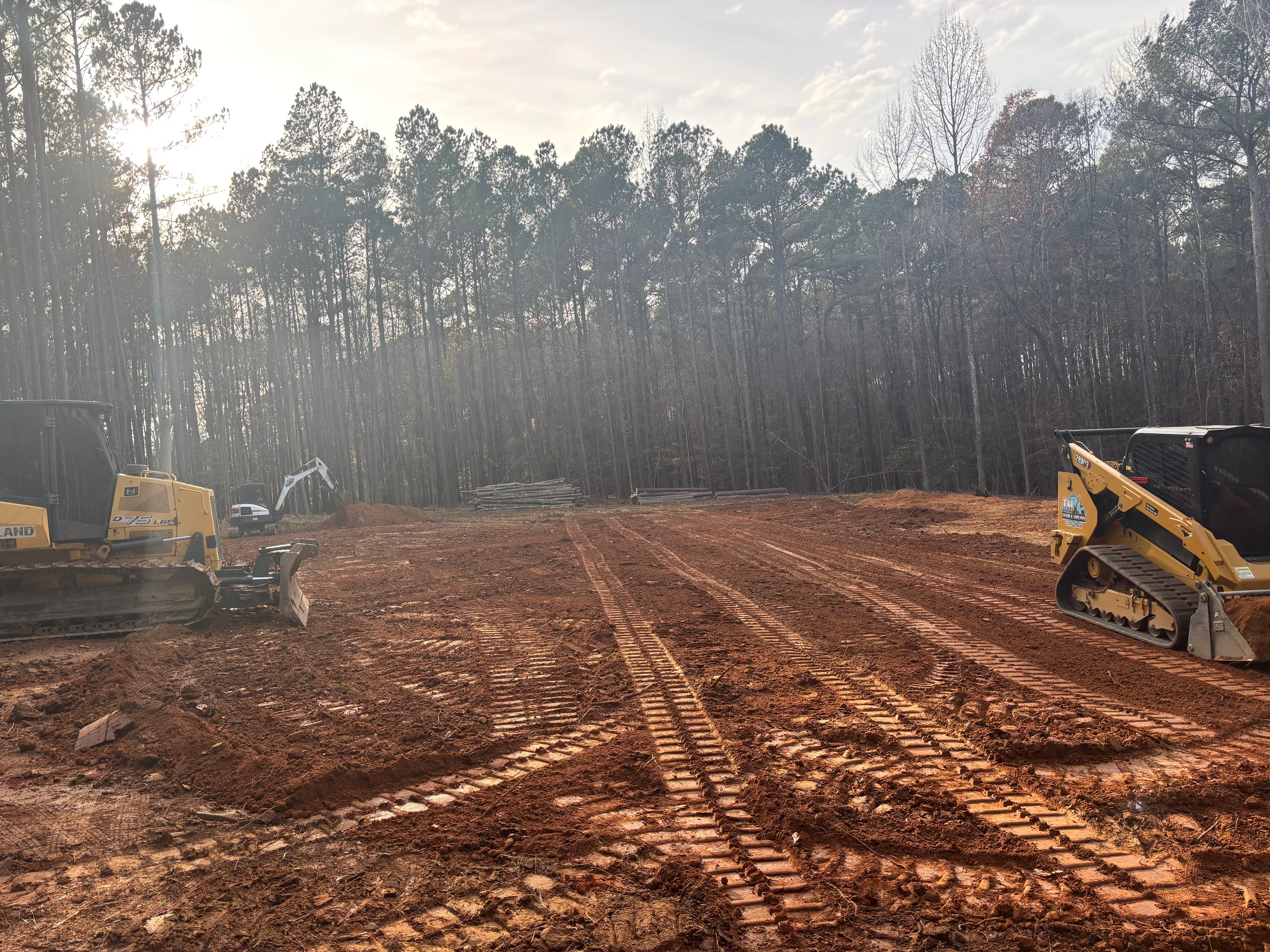 Two yellow tracked skid steers grading cleared land with pine forest backdrop