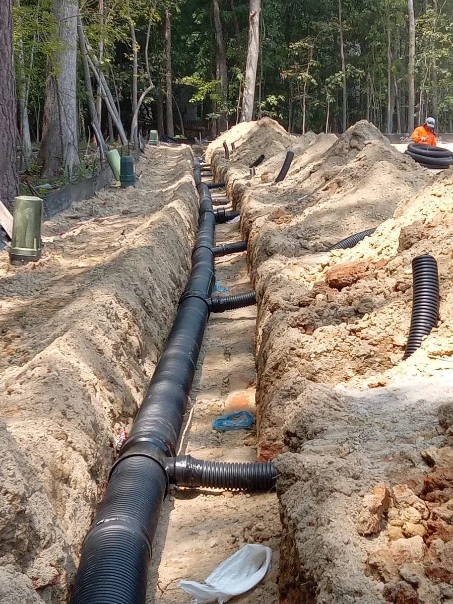 Black corrugated drainage pipes installed in parallel trenches with excavated soil and forest backdrop