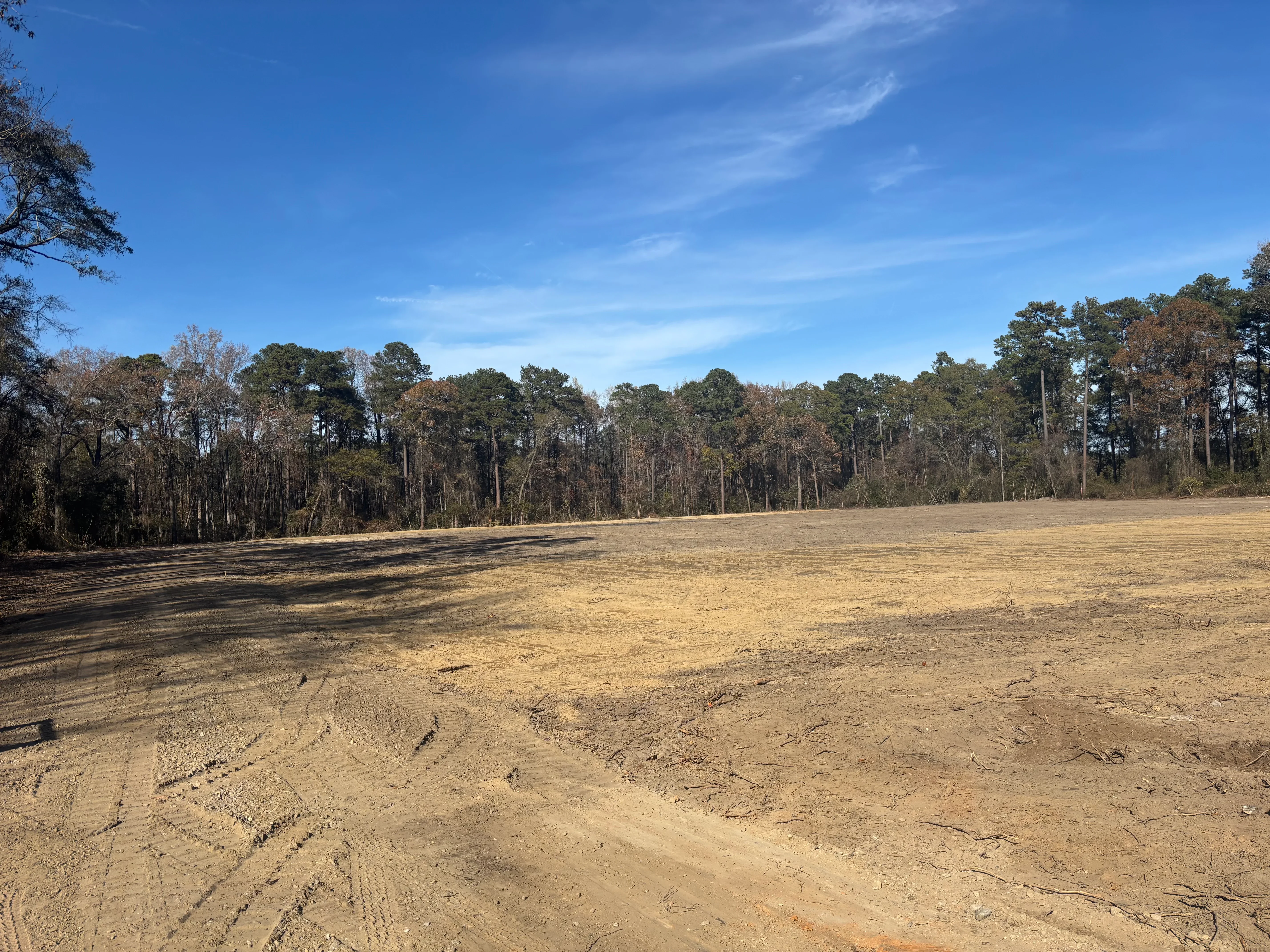 Cleared and graded dirt lot with tree line, showing excavation and site preparation work