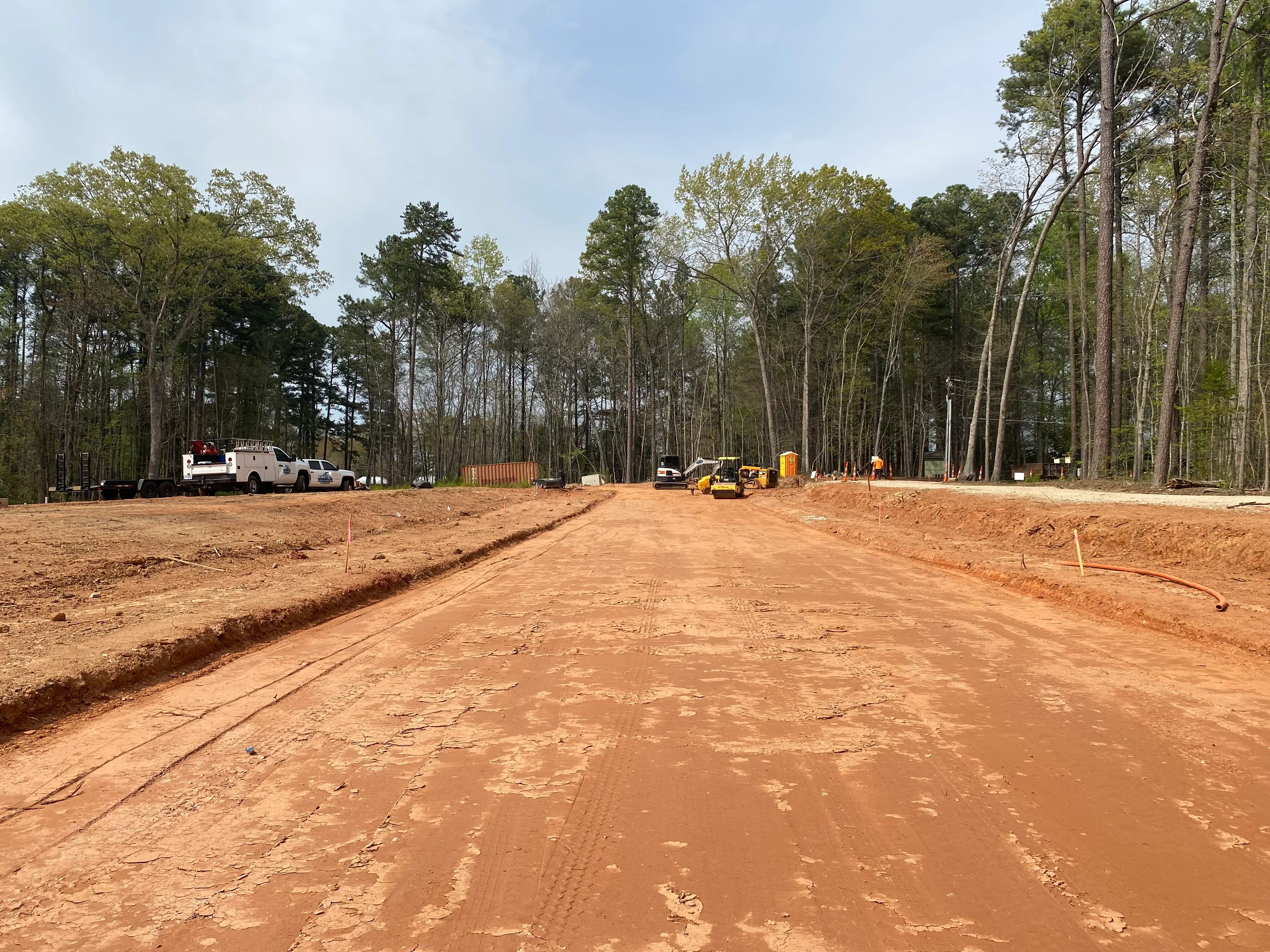 Excavated and graded red clay site with utility trenches, equipment, and cleared wooded perimeter