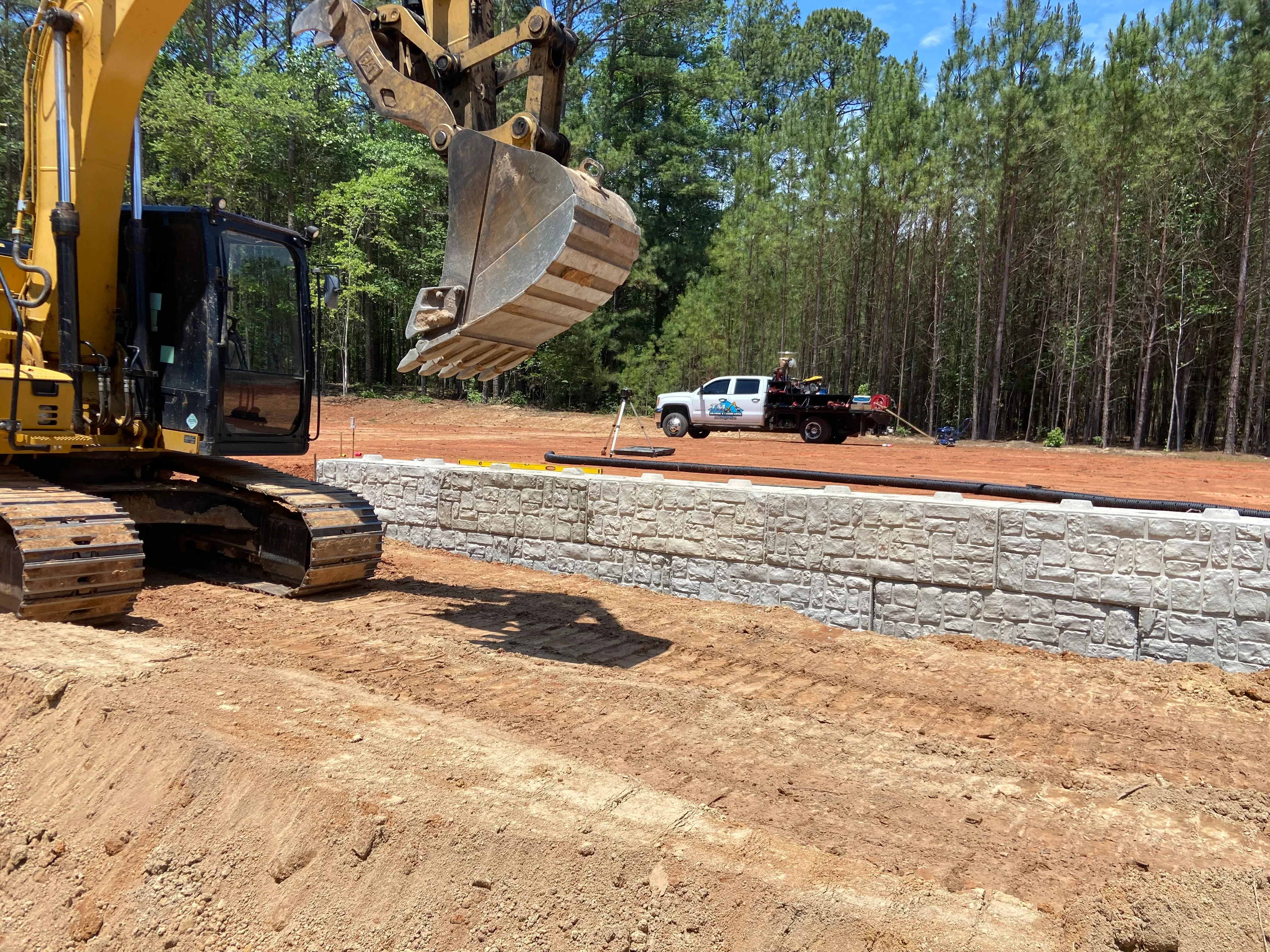 Excavator with bucket positioning soil near completed stone retaining wall on graded site
