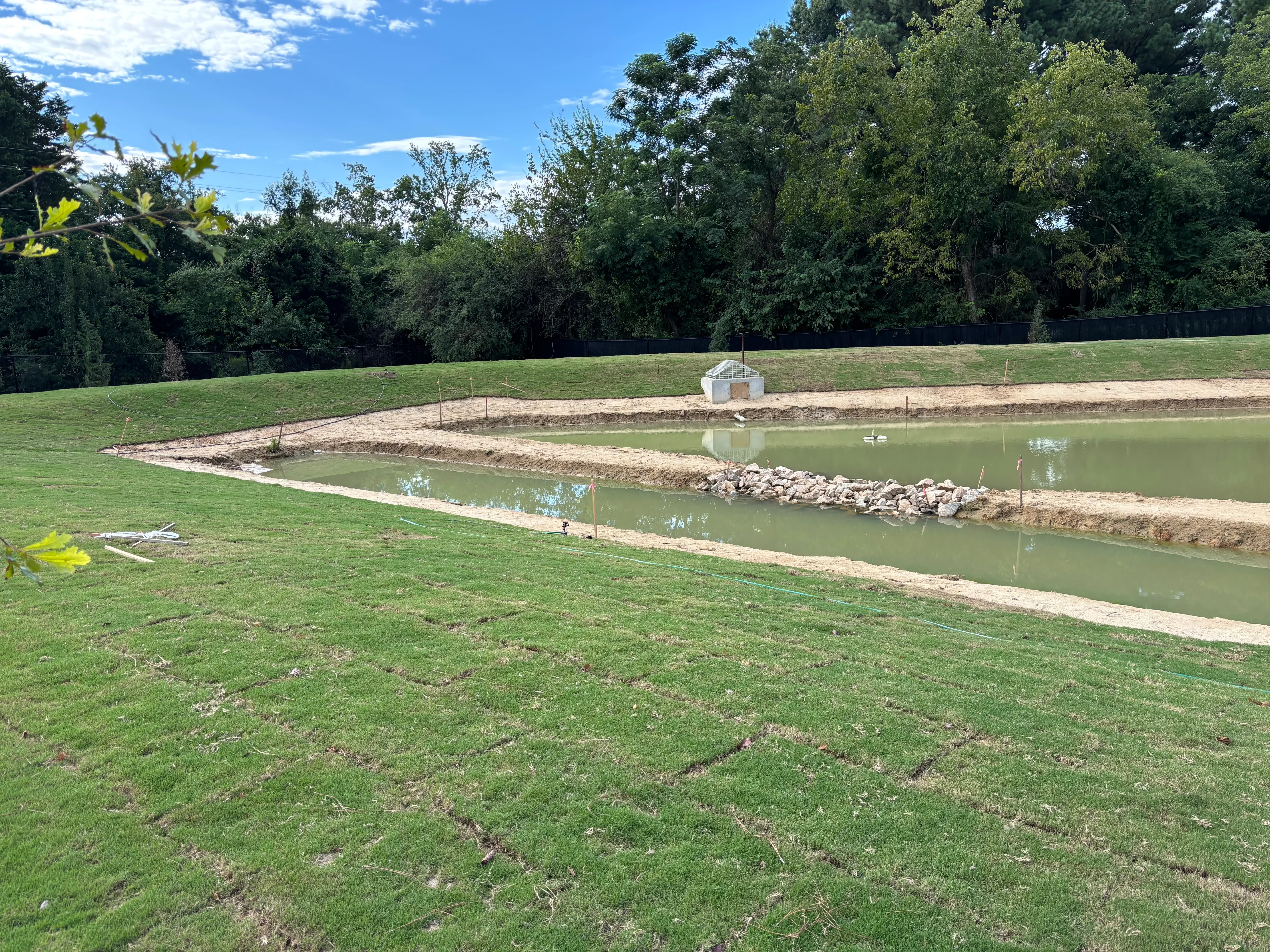 Excavated pond with rock riprap banks, cleared earthwork, and equipment shelter on landscaped property