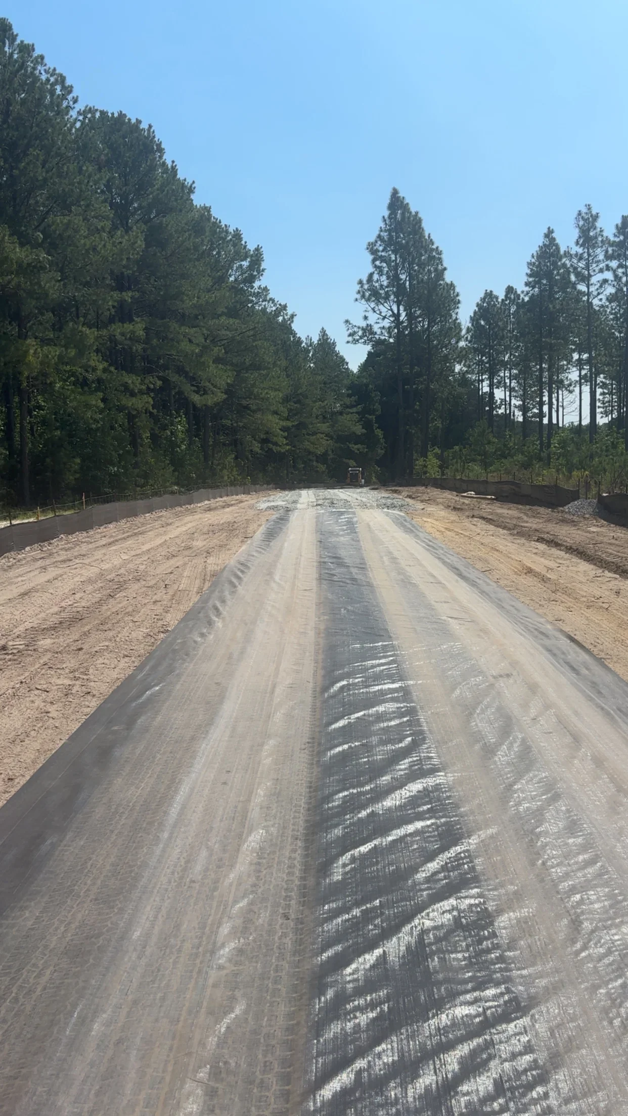 Graded driveway with geotextile fabric, compacted soil base, and metal fencing along tree-lined property