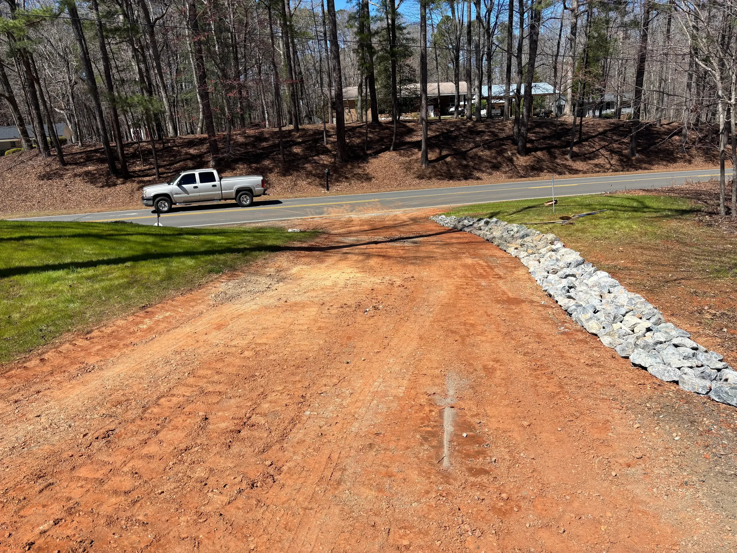 Excavated driveway base with red clay, stone erosion control border, and graded lawn transition