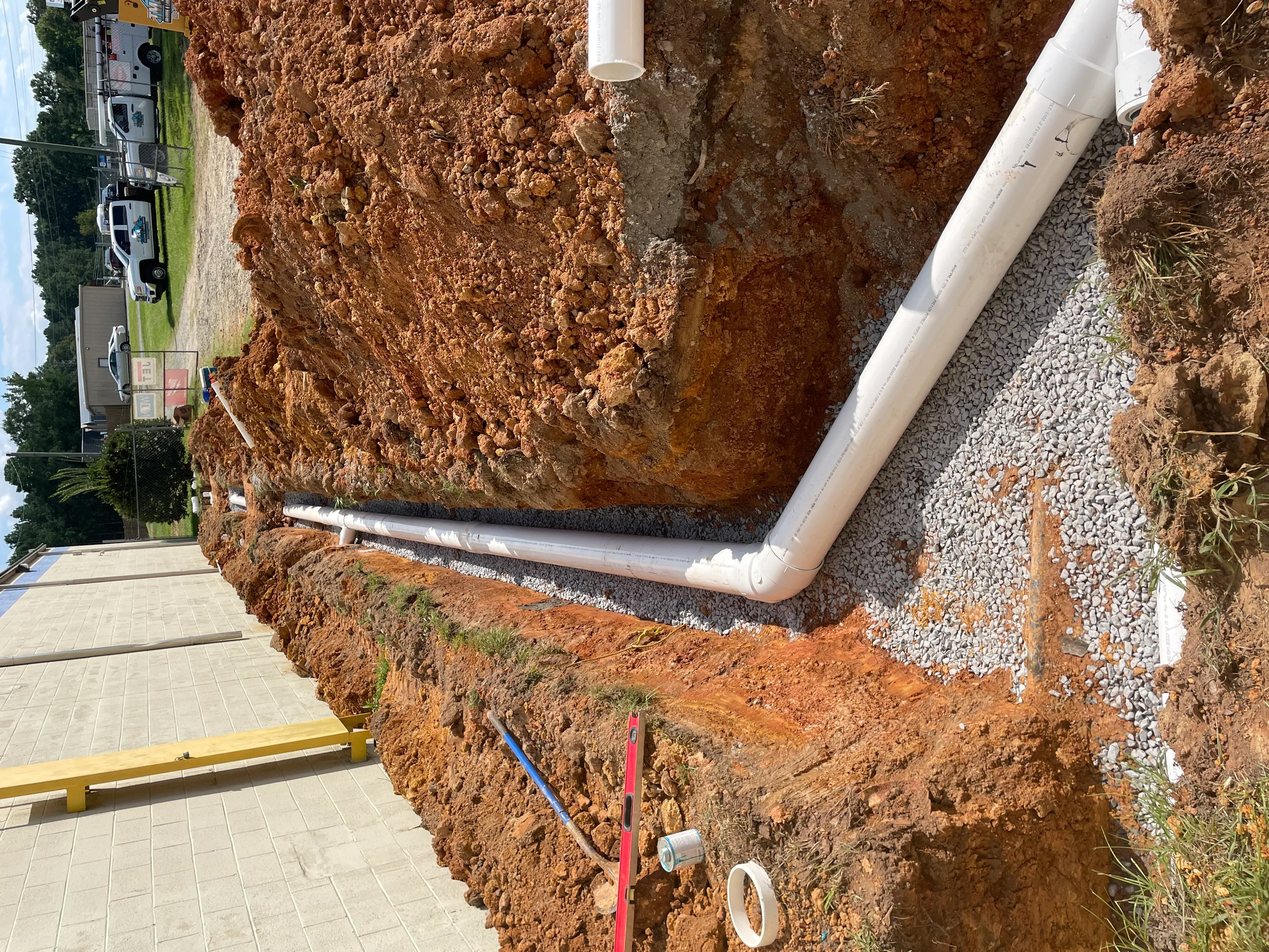 Excavated trench with white PVC drainage pipe, gravel base, and utility lines during installation